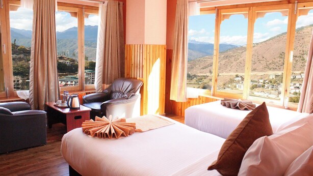 A hotel room with two beds, a chair, a table with a kettle, and a view of mountains. - Hotel Oro Villa (Thimphu)