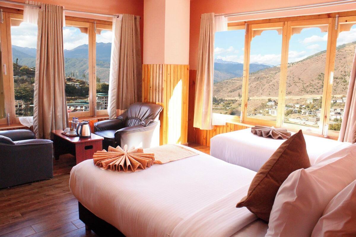 A hotel room with two beds, a chair, a table with a kettle, and a view of mountains.
