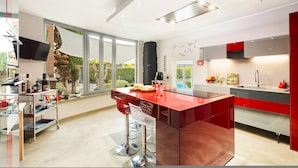 A modern kitchen with a central island, a dining area with bar stools, and a large window offering a view of the outdoors. - Villa Tiberio 6 2 Emma Villas (Genzano di Roma)