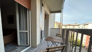 Een balkon met een houten bankje en een stoel met kussens, een betegelde vloer en uitzicht op omliggende gebouwen.