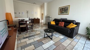 Een woonkamer met een zwarte leren bank, een eettafel met stoelen, een betegelde vloer, een radiator en een ingelijst kunstwerk aan de muur.
