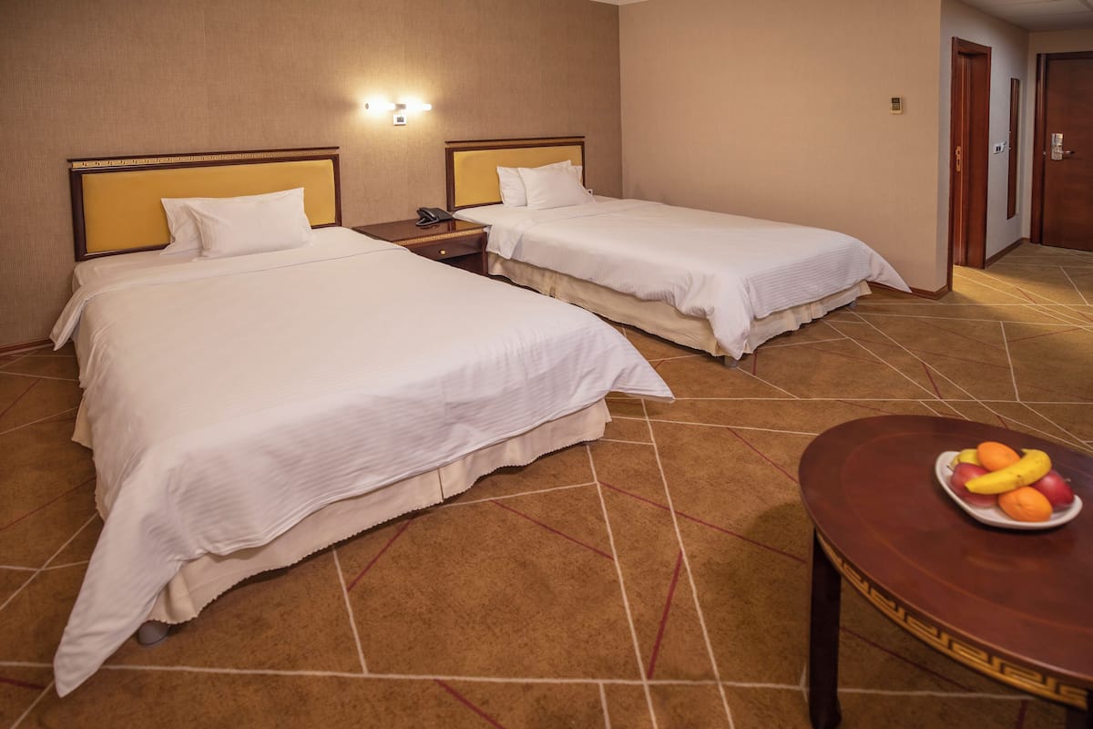 A hotel room with two beds, a bedside table with a phone, and a small table with fruit.