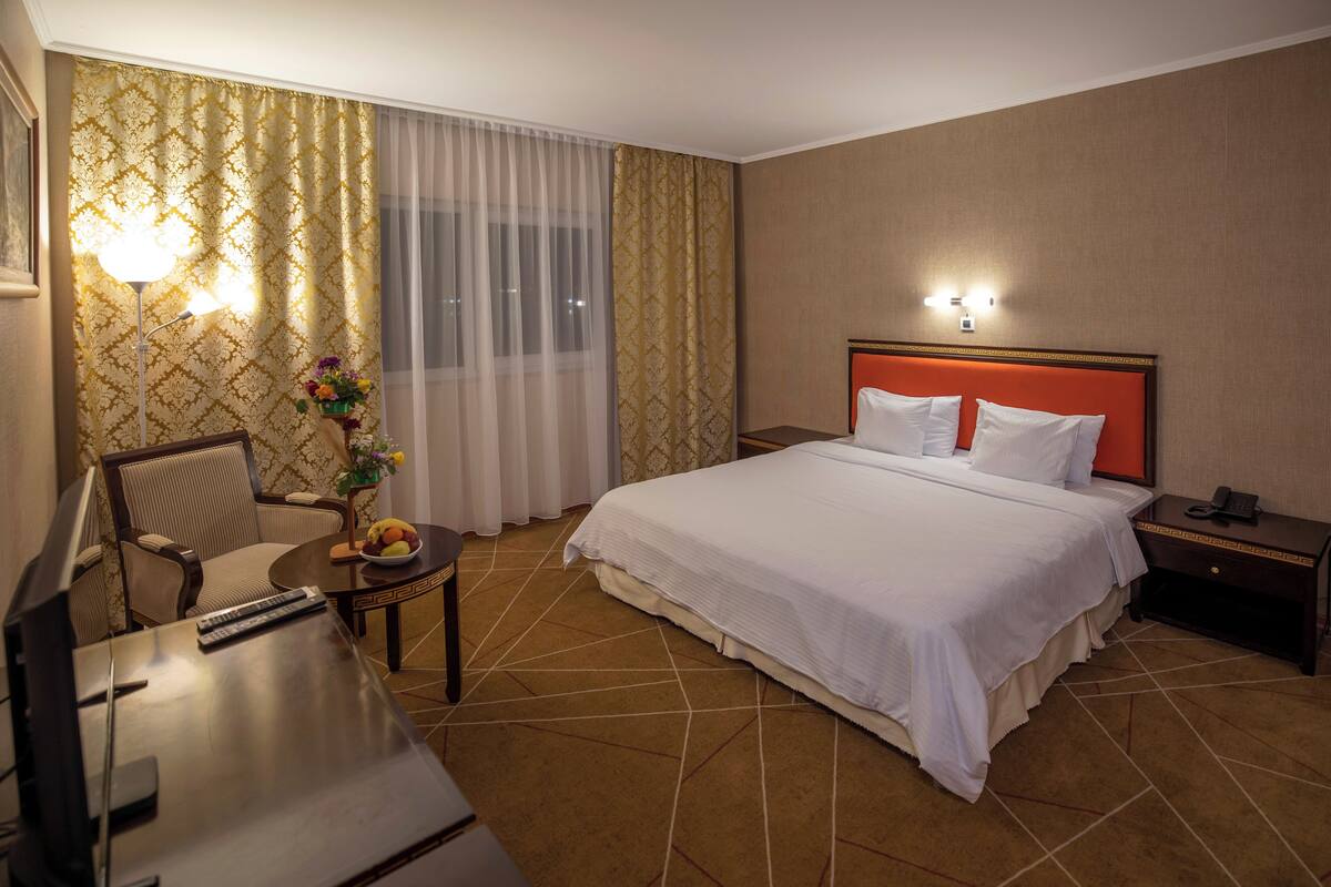 A hotel room with a bed, a desk with a chair, a small table with fruit, and a nightstand with a phone.