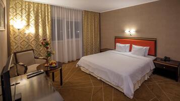 A hotel room with a bed, a desk with a chair, a small table with fruit, and a nightstand with a phone.