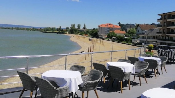 A terrace with wicker chairs and tables overlooking a beach and a calm body of water.