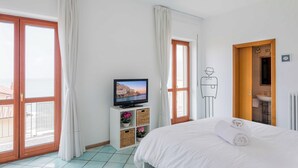 Une chambre d’hôtel avec un lit, un téléviseur posé sur un meuble, une salle de bain avec un lavabo et un miroir, et de grandes fenêtres avec des rideaux.