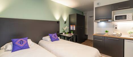 A hotel room with two beds, a small kitchenette, and a desk with a chair.
