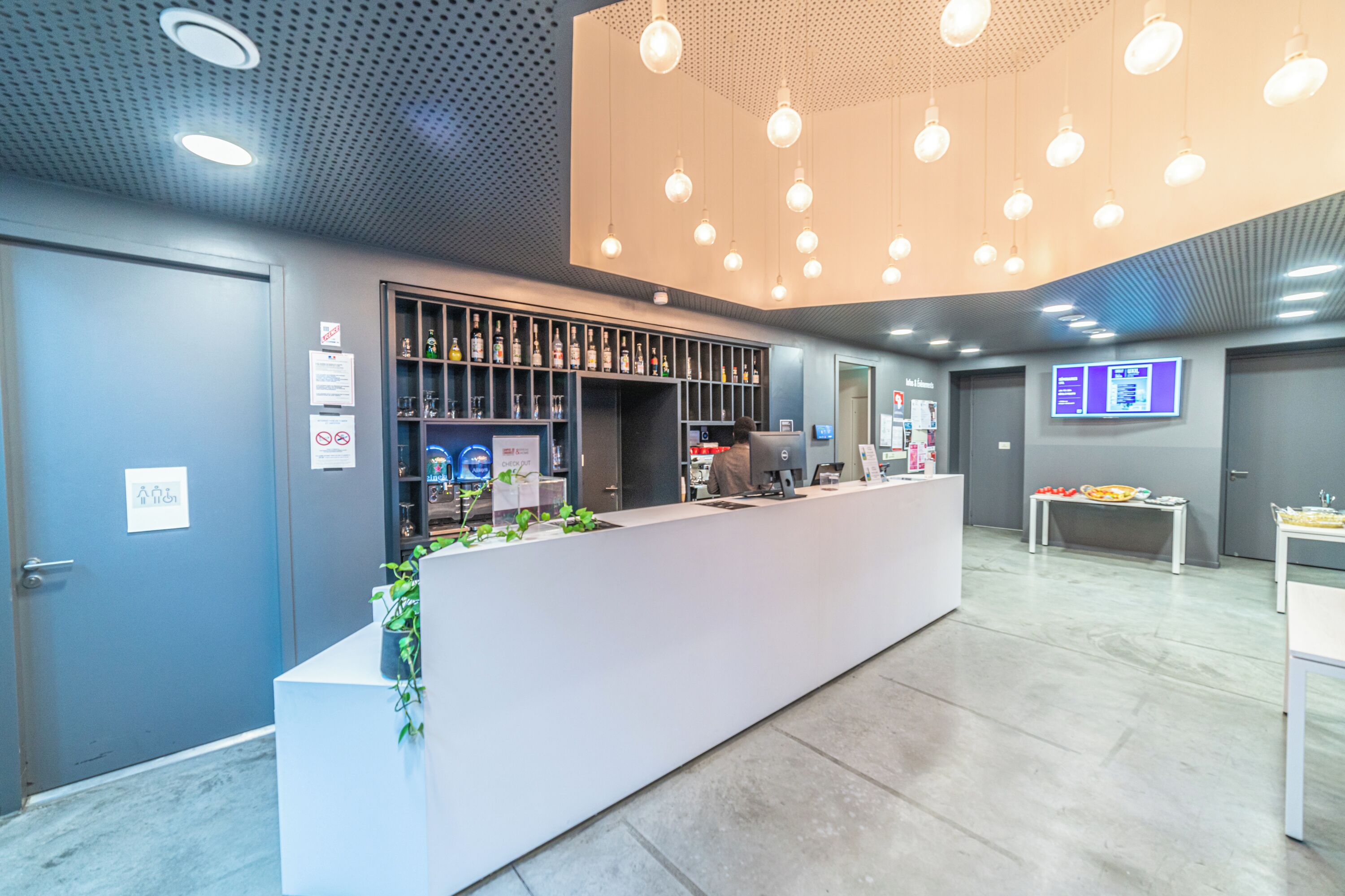 A modern reception area with a white counter, a plant, and a bar with shelves of bottles.