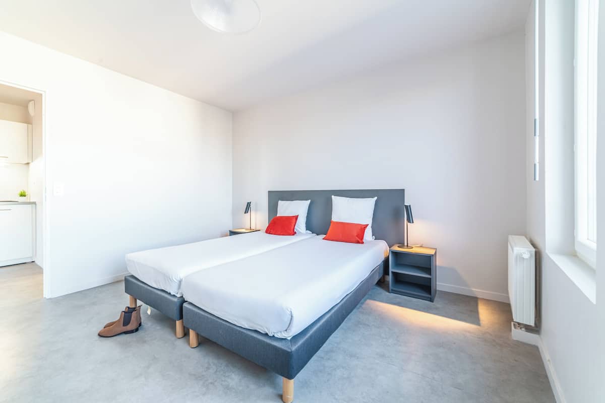 A modern bedroom with a large bed, bedside tables, a radiator, and a view of the adjacent kitchen.