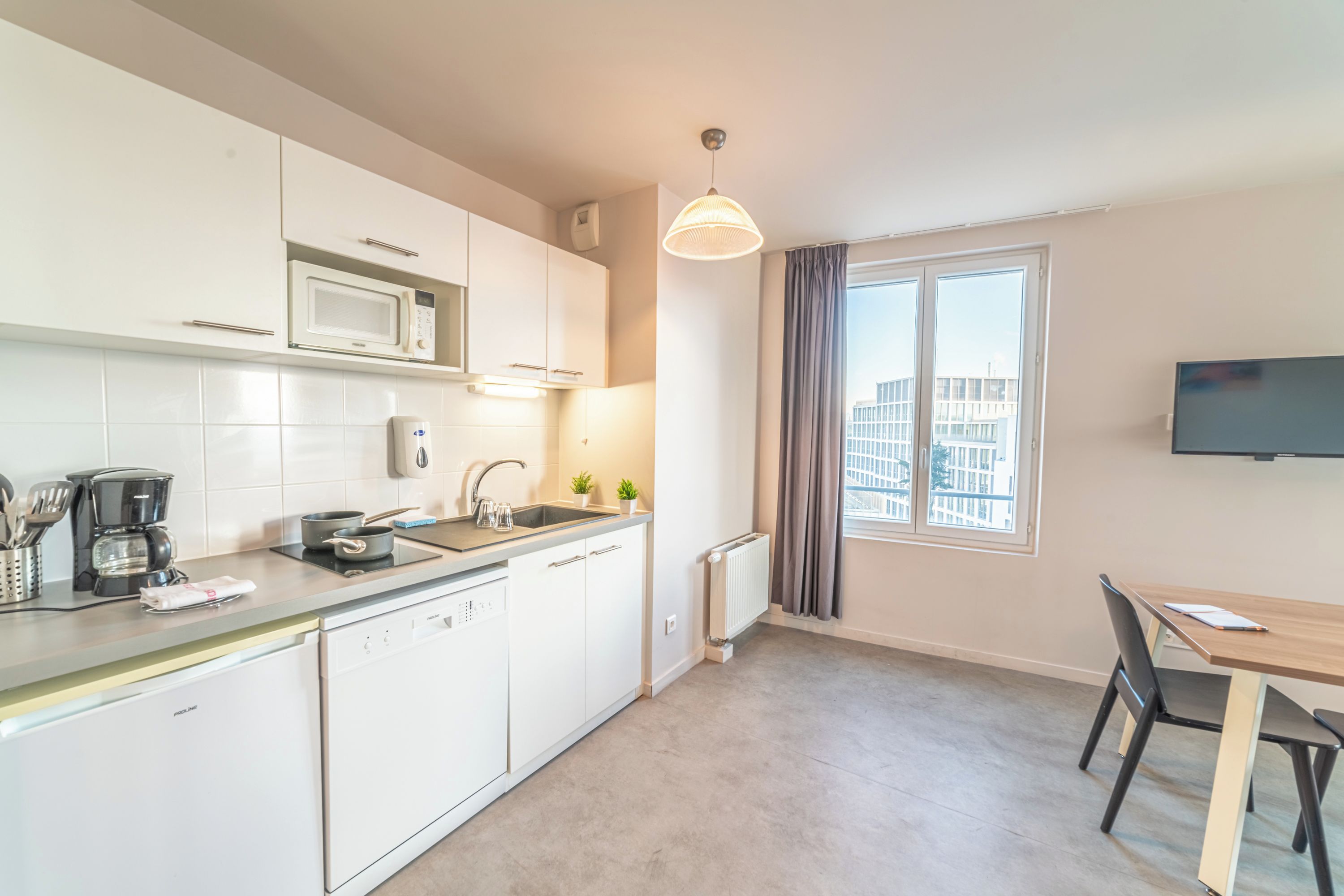 Una cocina moderna con gabinetes blancos, microondas integrado, cafetera y pileta. Hay un comedor con mesa y sillas, y una ventana con cortinas.