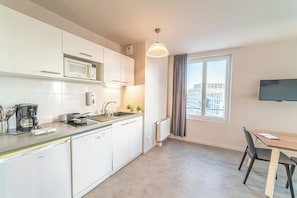 Una cocina moderna con gabinetes blancos, microondas integrado, cafetera y pileta. Hay un comedor con mesa y sillas, y una ventana con cortinas.