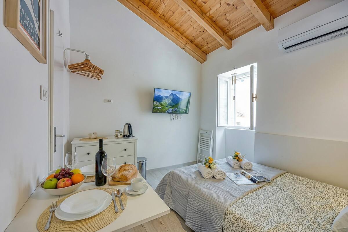 A bedroom with a bed, a bedside table with a bowl of fruit, a bottle, and glasses, a television on the wall, and a window with a view.