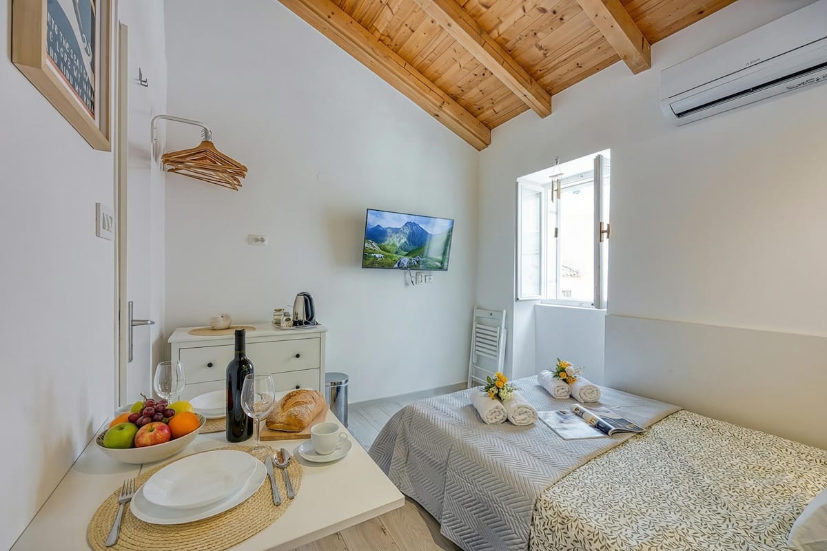 A bedroom with a bed, a bedside table with a bowl of fruit, a bottle, and glasses, a television on the wall, and a window with a view.
