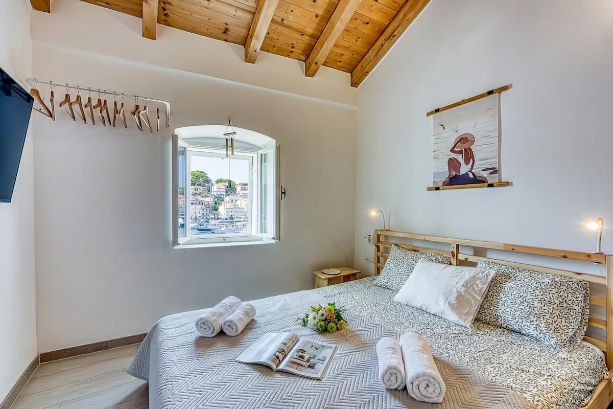A bedroom with a bed, a window with a view, a hanging art piece, and a wooden ceiling.