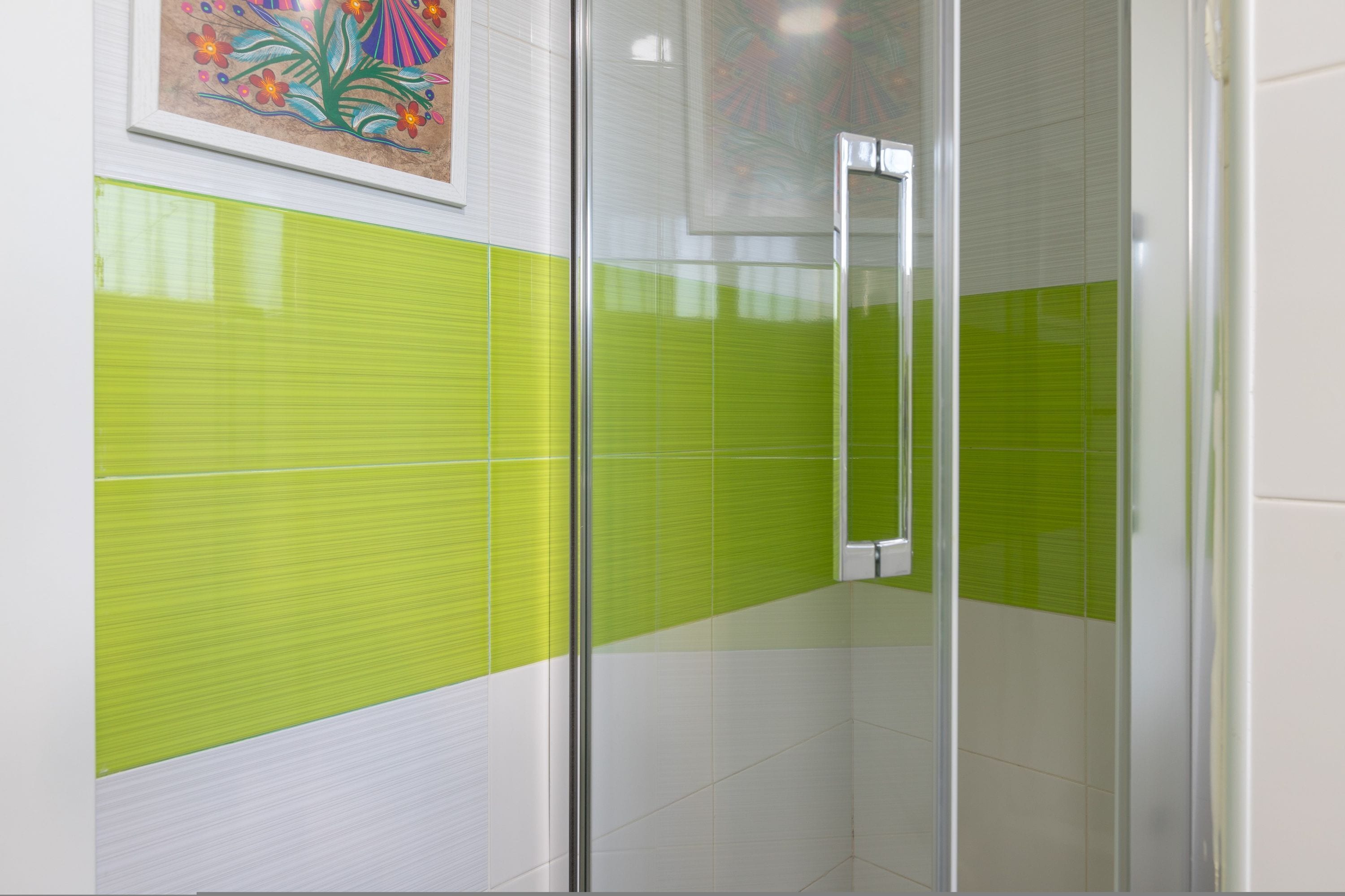 A bathroom with green and white tiled walls, a glass shower enclosure, and a framed artwork on the wall.