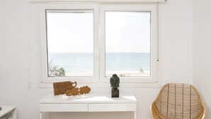 Ein Zimmer mit einem weißen Schreibtisch, einem Korbstuhl und Meerblick durch zwei Fenster.