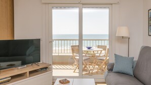 Ein modernes Wohnzimmer mit Flachbildfernseher, Couchtisch und einer gepolsterten Couch. Ein Balkon mit Tisch und Stühlen bietet Blick auf das Meer.