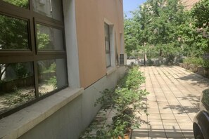 Fachada de un edificio con un patio empedrado, una ventana con persianas y un camino bordeado de árboles.