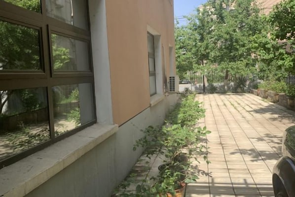 Fachada de un edificio con un patio empedrado, una ventana con persianas y un camino bordeado de árboles.