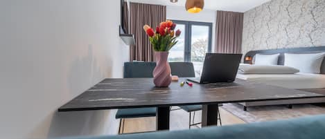 A modern hotel room with a bed, a laptop on the bedside table, a vase of flowers, and a view of the city through the window.