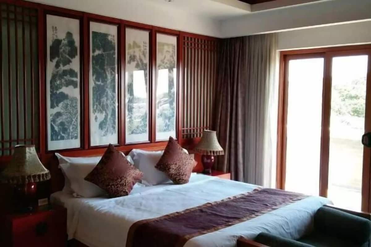Une chambre d’hôtel avec un lit, des oreillers, une table de chevet, une lampe, une fenêtre avec des rideaux et des œuvres d’art au mur.