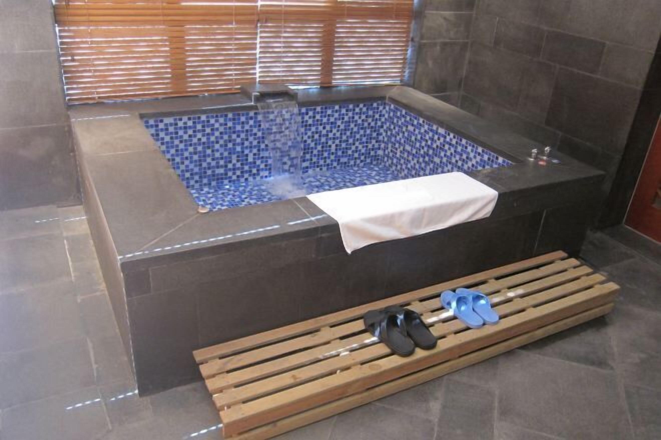 A bathroom with a tiled hot tub, a bench with shoes, and a window with blinds.