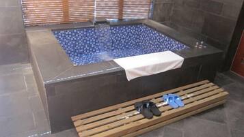 A bathroom with a tiled hot tub, a bench with shoes, and a window with blinds.