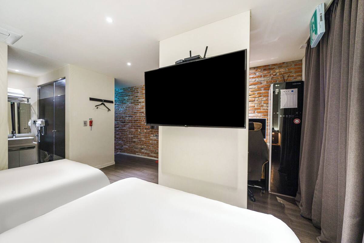 Ein Hotelzimmer mit Flachbildfernseher an der Wand, einem Bett, einem Schreibtisch mit Stuhl, einem Spiegel und einer Wand mit Ziegeloptik.