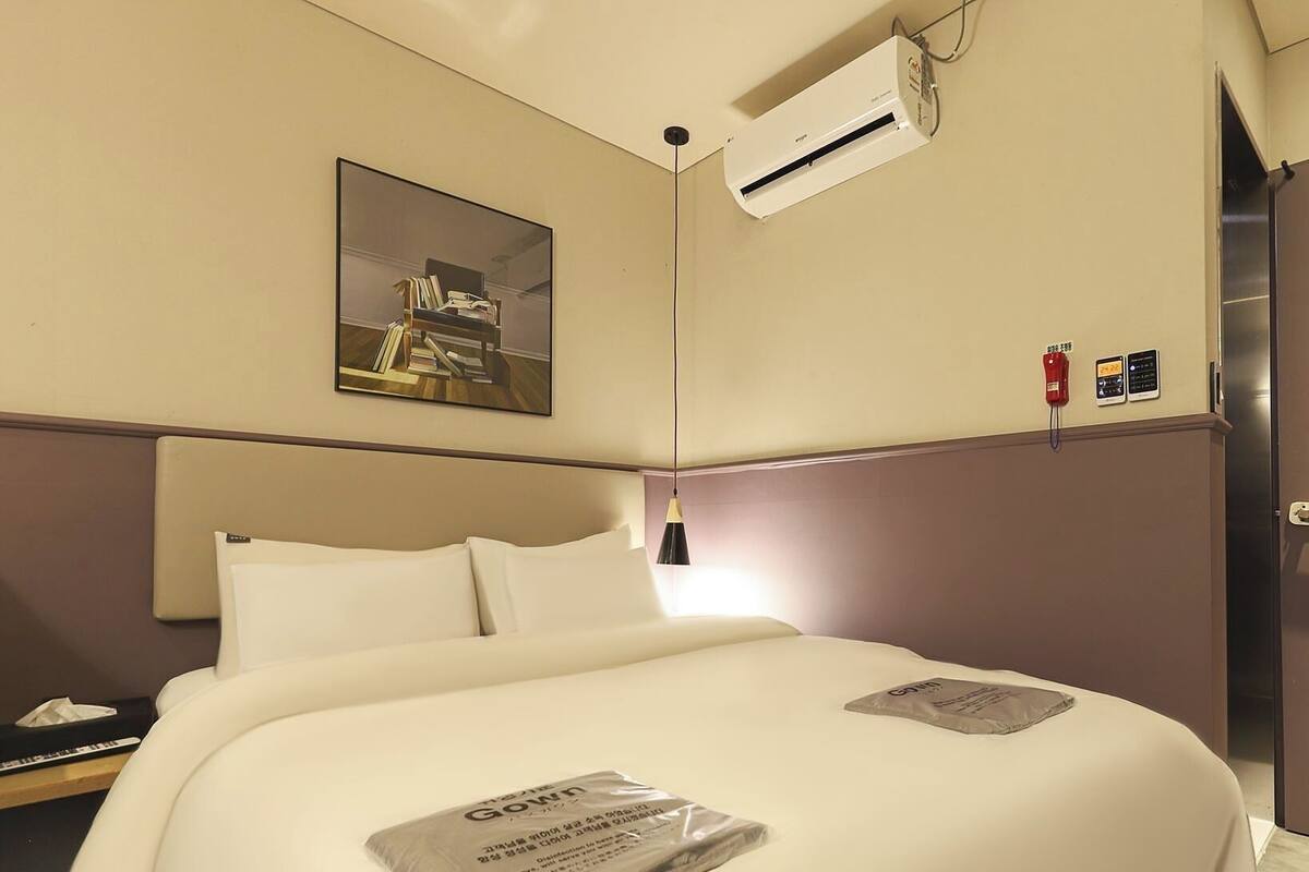 Ein Hotelzimmer mit Bett, Nachttisch, Lampe, Klimaanlage, einem Gemälde und einem Rauchmelder.