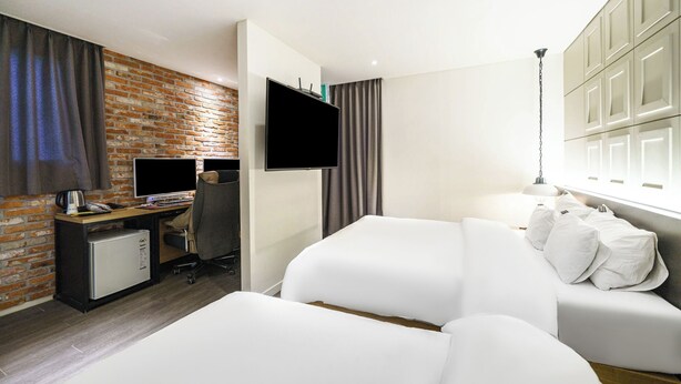 Une chambre d’hôtel moderne avec un mur d’accent en briques, un téléviseur à écran plat, un bureau avec un ordinateur, un petit réfrigérateur et un canapé blanc.