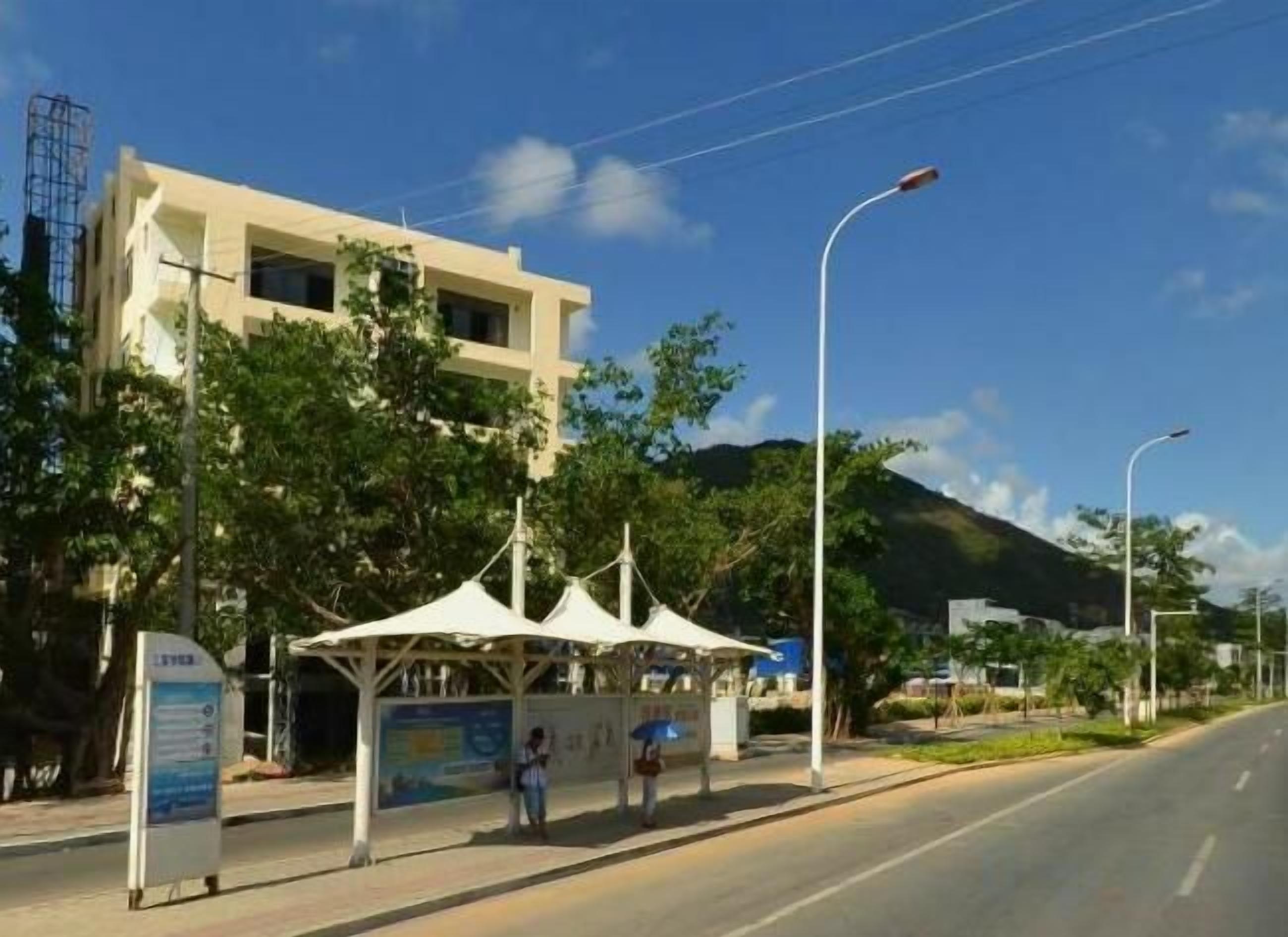 A bus stop with a shelter, a signboard, and a road with a mountain in the background.