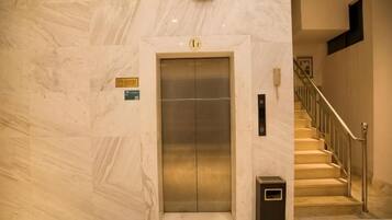 An elevator with a sign indicating it is the first floor, a trash bin beside it, and a staircase with a handrail leading to an upper level.
