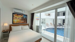 A hotel room with a bed, a nightstand, a lamp, a painting of a sunset, and a view of a pool area with lounge chairs. - B14 - 2 Bedroom Serviced Apt with Pool Access, Karon Beach (Tambon Karon)