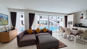 A modern living room with a sofa, ottoman, dining area, kitchen, and a pool view. - B14 - 2 Bedroom Serviced Apt with Pool Access, Karon Beach (Tambon Karon)