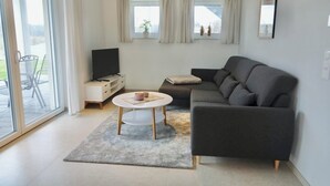 Ett modernt vardagsrum med en grå soffa, ett litet vitt bord och en tv på ett vitt skåp. Det finns en skjutdörr i glas som leder ut till ett utomhusområde med stolar.