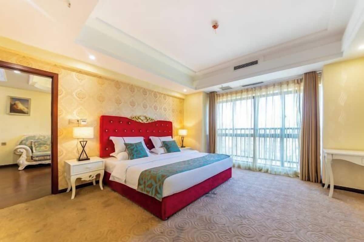 Une chambre d’hôtel avec un lit rouge, une table de chevet, une lampe, une fenêtre avec des rideaux et une chaise.
