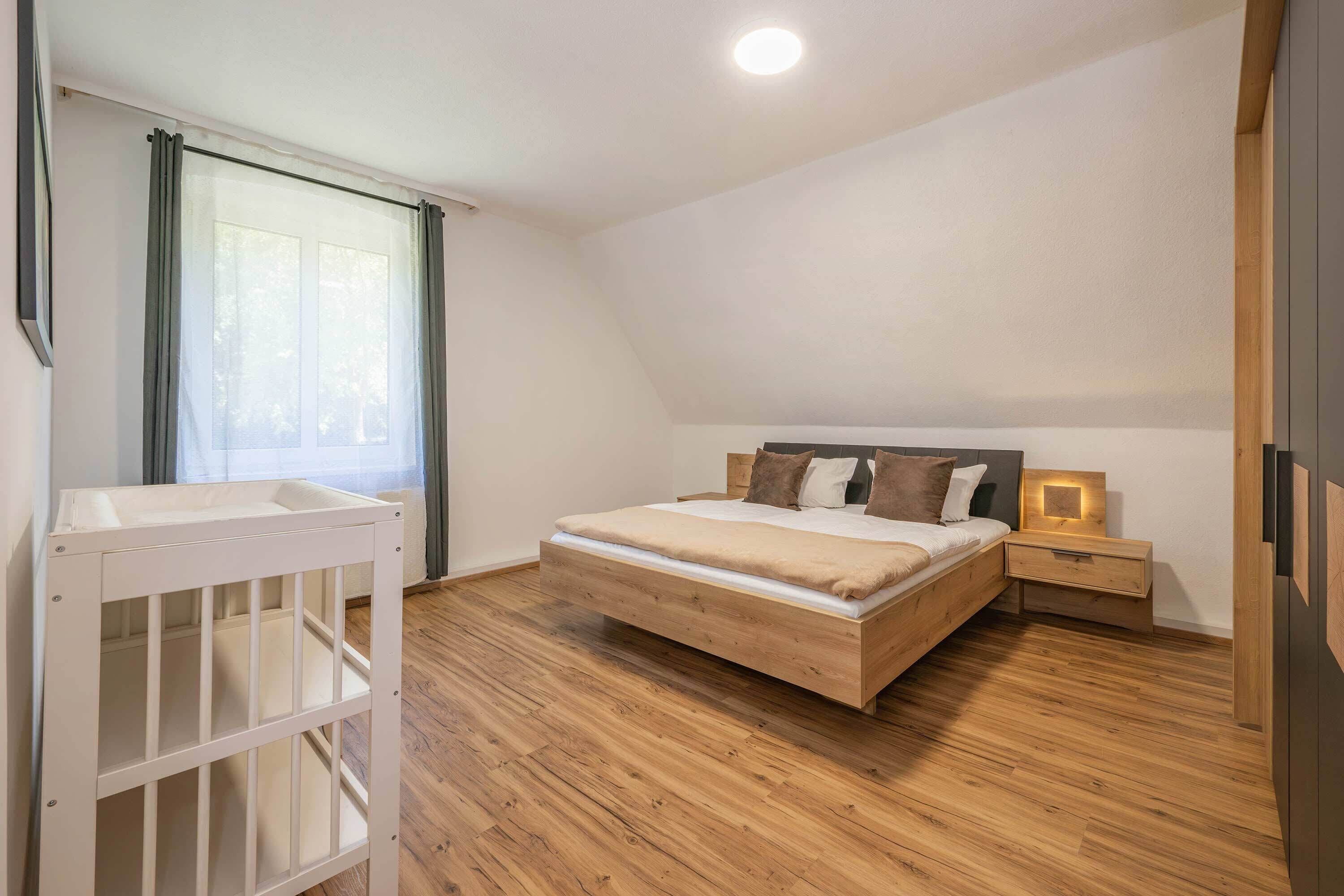 A bedroom with a wooden bed, a nightstand, a crib, and a window with curtains.