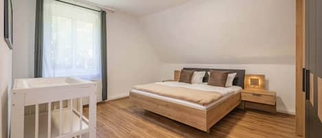 A bedroom with a wooden bed, a nightstand, a crib, and a window with curtains.