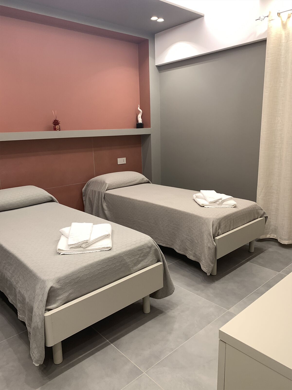 Una habitación de hotel con dos camas, un estante pequeño con objetos decorativos y una cortina.