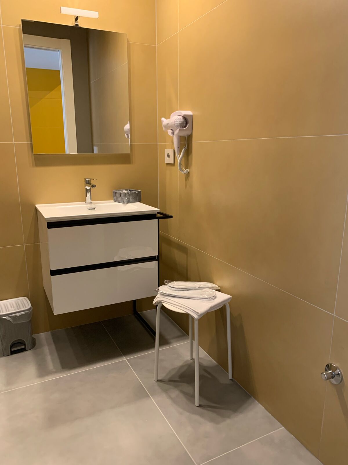 A modern bathroom with a white vanity, a mirror, a hairdryer, and a small stool.
