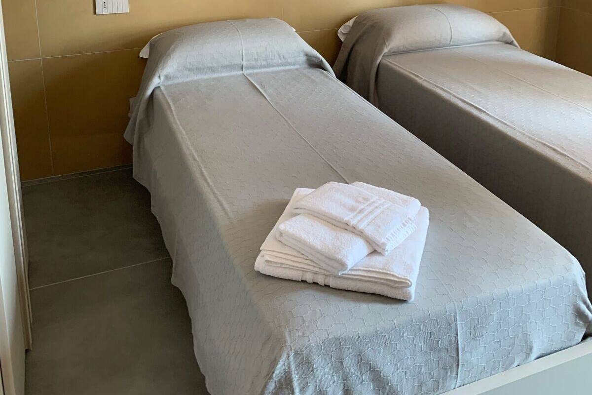 A room with two single beds, each with a grey bedspread and white pillows. There is a folded white towel on the bed in the foreground. The walls are a light brown color, and there is a white shelf above the beds.