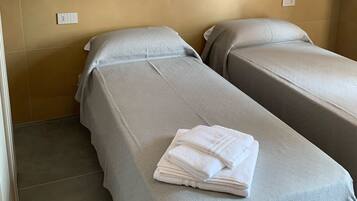 Una camera con due letti singoli, ognuno con copripiumino grigio e cuscini bianchi. C'è un asciugamano bianco piegato sul letto in primo piano. Le pareti sono di un colore marrone chiaro, e c'è uno scaffale bianco sopra i letti.