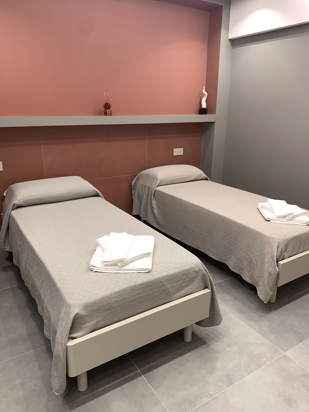 Zwei Einzelbetten mit grauer Bettwäsche und weißen Handtüchern, eine rote Wand und ein grauer Boden in einem Hotelzimmer.