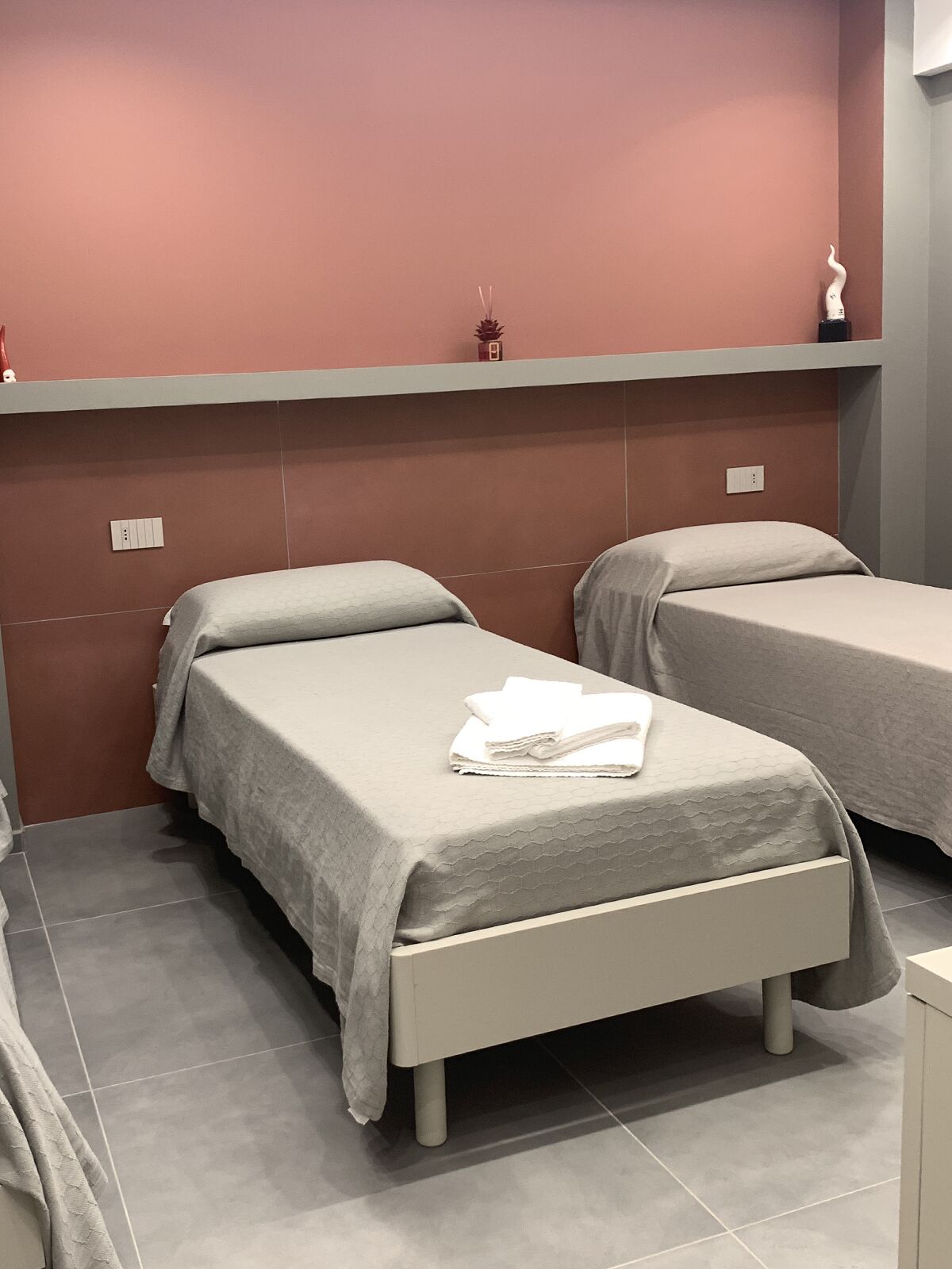 A hotel room with two single beds, a small shelf with decorative items, and a tiled floor.
