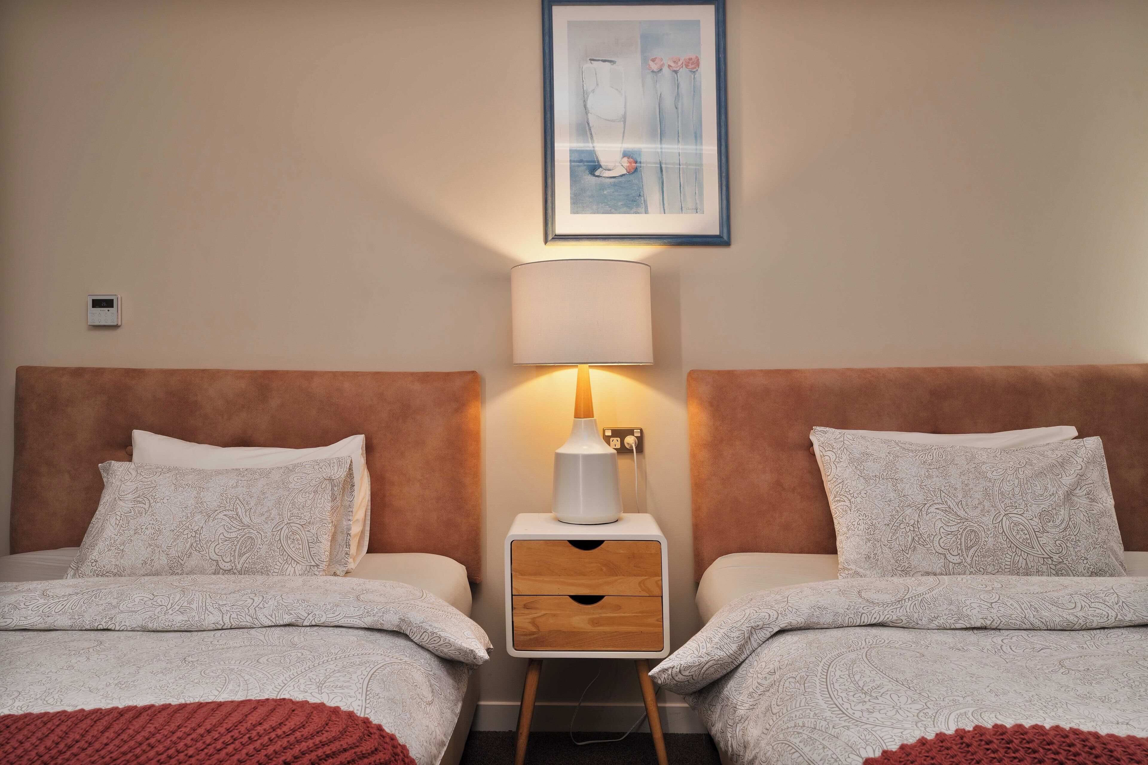 A hotel room with two beds, a nightstand with a lamp, a framed artwork on the wall, and a red knitted blanket on the floor.