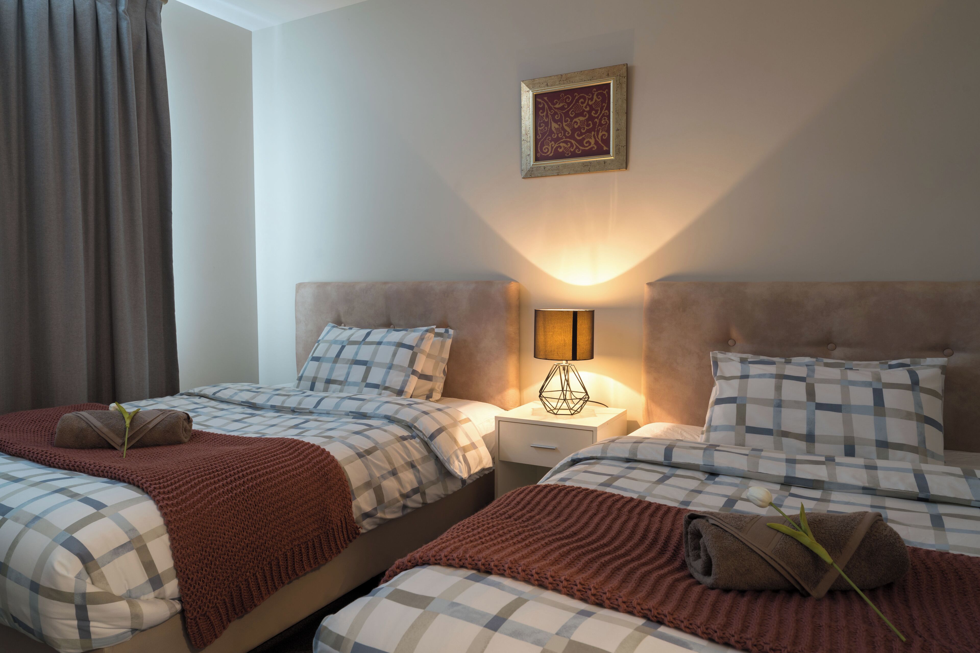 A hotel room with two beds, each with a checkered bedspread, a brown blanket, and a rolled towel. There is a nightstand with a lamp between the beds.