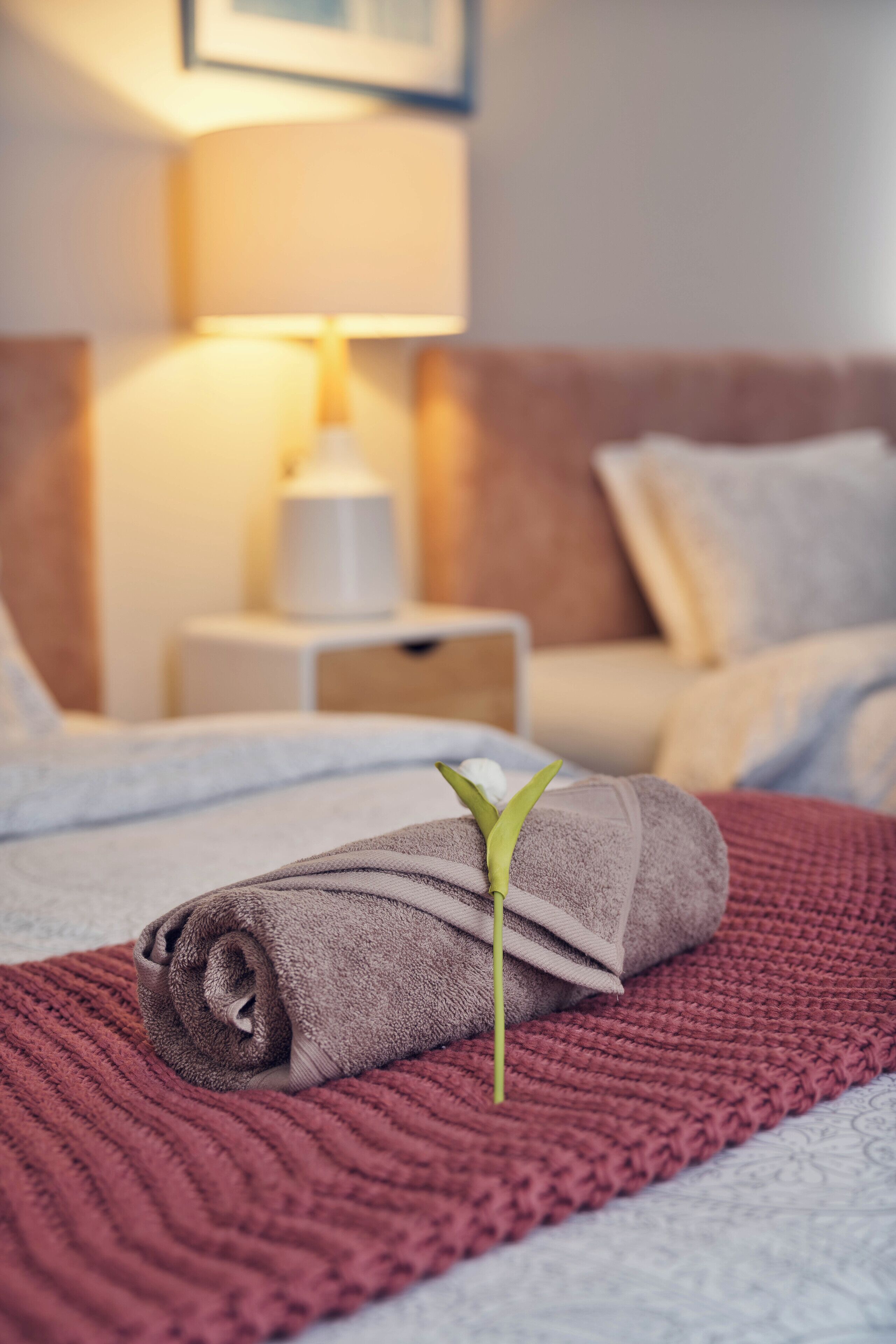 A neatly made bed with a rolled towel and a green plant, a bedside lamp, and a nightstand with a lamp.