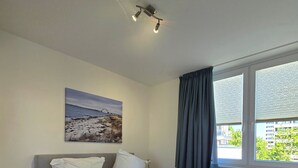 Un dormitorio con cama, mesita de noche, ventana con cortinas y un cuadro en la pared.