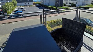 Un balcón con una silla de mimbre, una mesa y vistas a un estacionamiento y edificios.