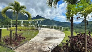 Un sendero curvo de hormigón que conduce a una estructura de pérgola blanca, rodeada de plantas tropicales y palmeras, con montañas al fondo.